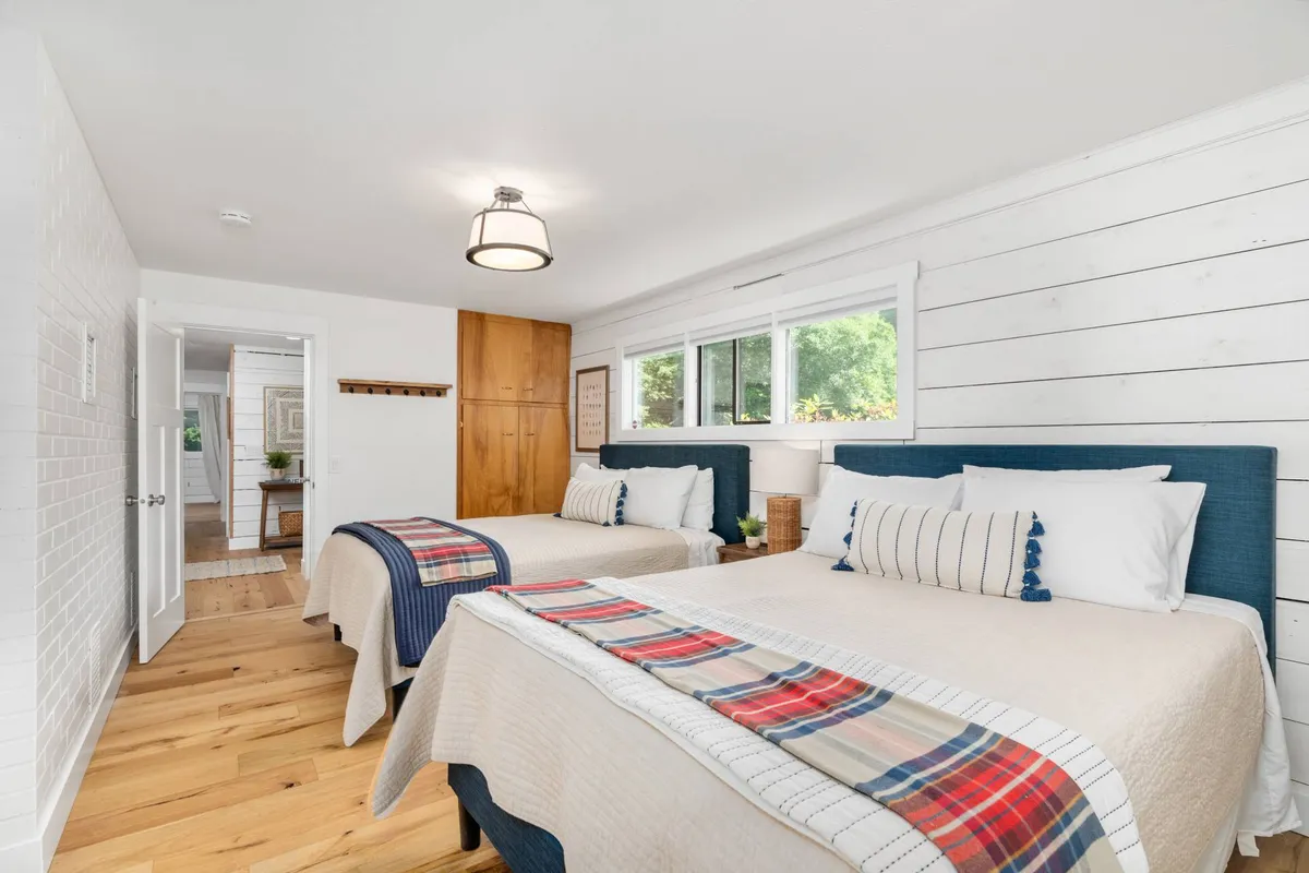 A bedroom with two beds, each decorated with cozy throws, and large windows that fill the room with natural light. The wooden closet and warm tones create a welcoming atmosphere, ideal for siblings or friends sharing a space.