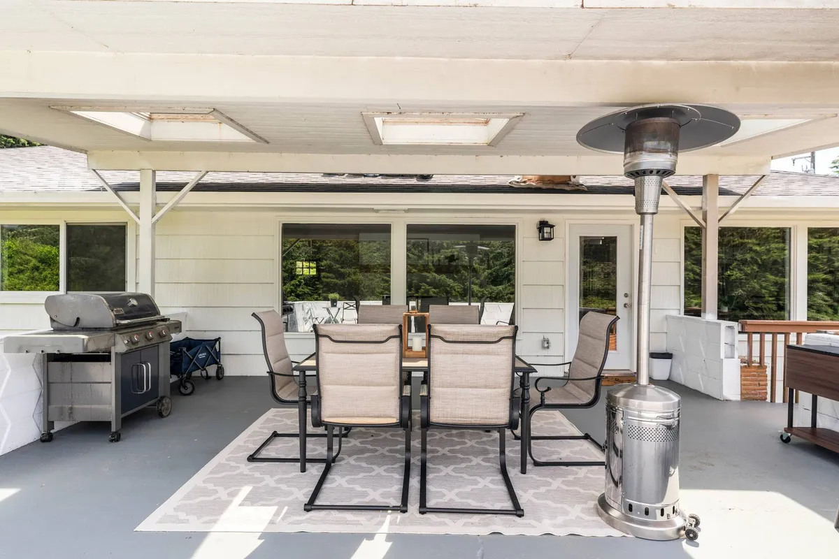 An inviting outdoor patio features a dining table set for gatherings, a grill, patio heater, and a cozy rug. The setting is designed for outdoor dining, socializing, or relaxing after a day on the water, and hints at the communal aspect of the lakeside retreat.