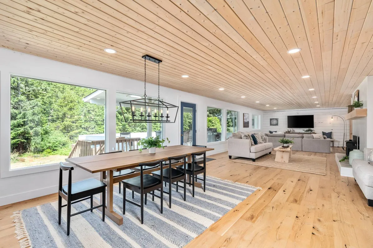 A warm, welcoming space showcasing a dining table adjacent to a sofa seating area, all bathed in light from large windows. The wooden ceiling and views of the outdoors add to the inviting, communal vibe. The space is clearly designed for gatherings, meals, and relaxed conversation.