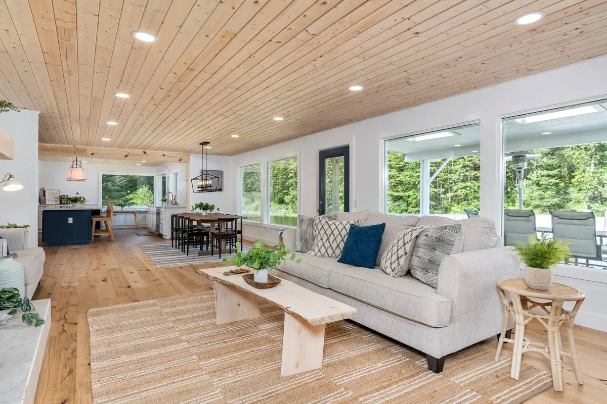 A bright, open-plan area with a sofa and dining table beneath a wooden ceiling, surrounded by large windows that frame views of the outdoors. The space invites both casual dining and relaxing while staying visually connected to the natural setting outside.