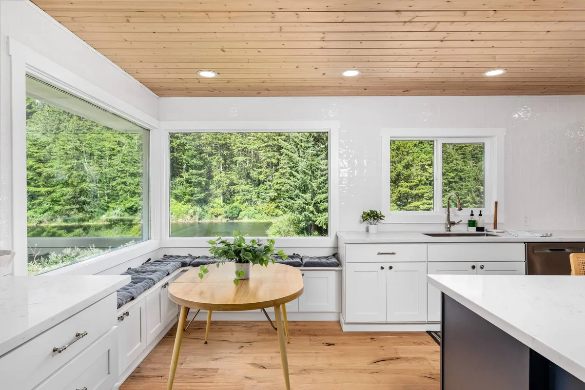 A calm and cozy dining area with large windows framing lush greenery outside, a round wooden table, and white cabinetry. The scene exudes tranquility while highlighting the beautiful natural surroundings visible from inside.