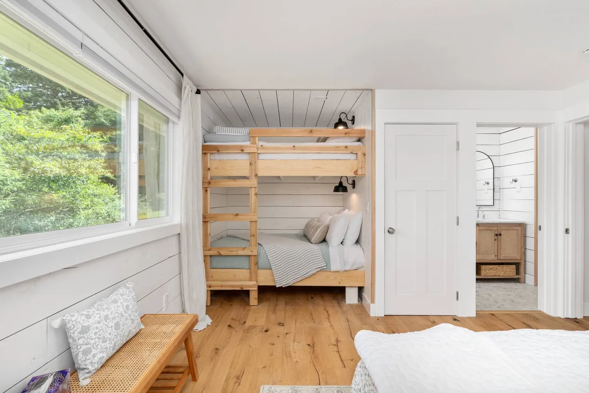 A cheerful bunk room featuring a sturdy bunk bed, large window, wood flooring, and a decorative bench. The daylight streaming in makes the space ideal for family bonding and offers a playful yet restful retreat for kids.