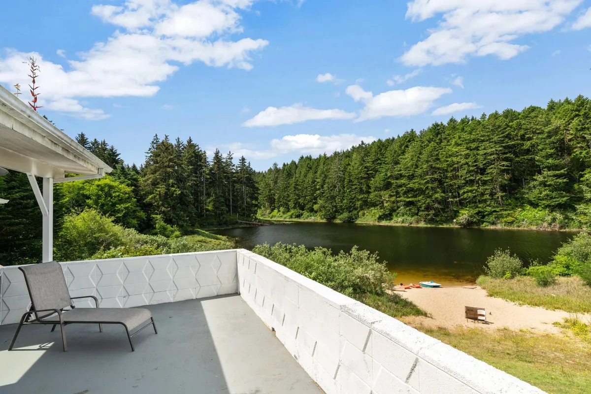 A tranquil view of the lake with surrounding forest, a sandy beach, and a deck chair for relaxing. The peaceful mood evokes early morning or evening moments by the water, ideal for swimming, relaxing, or watching sunsets.