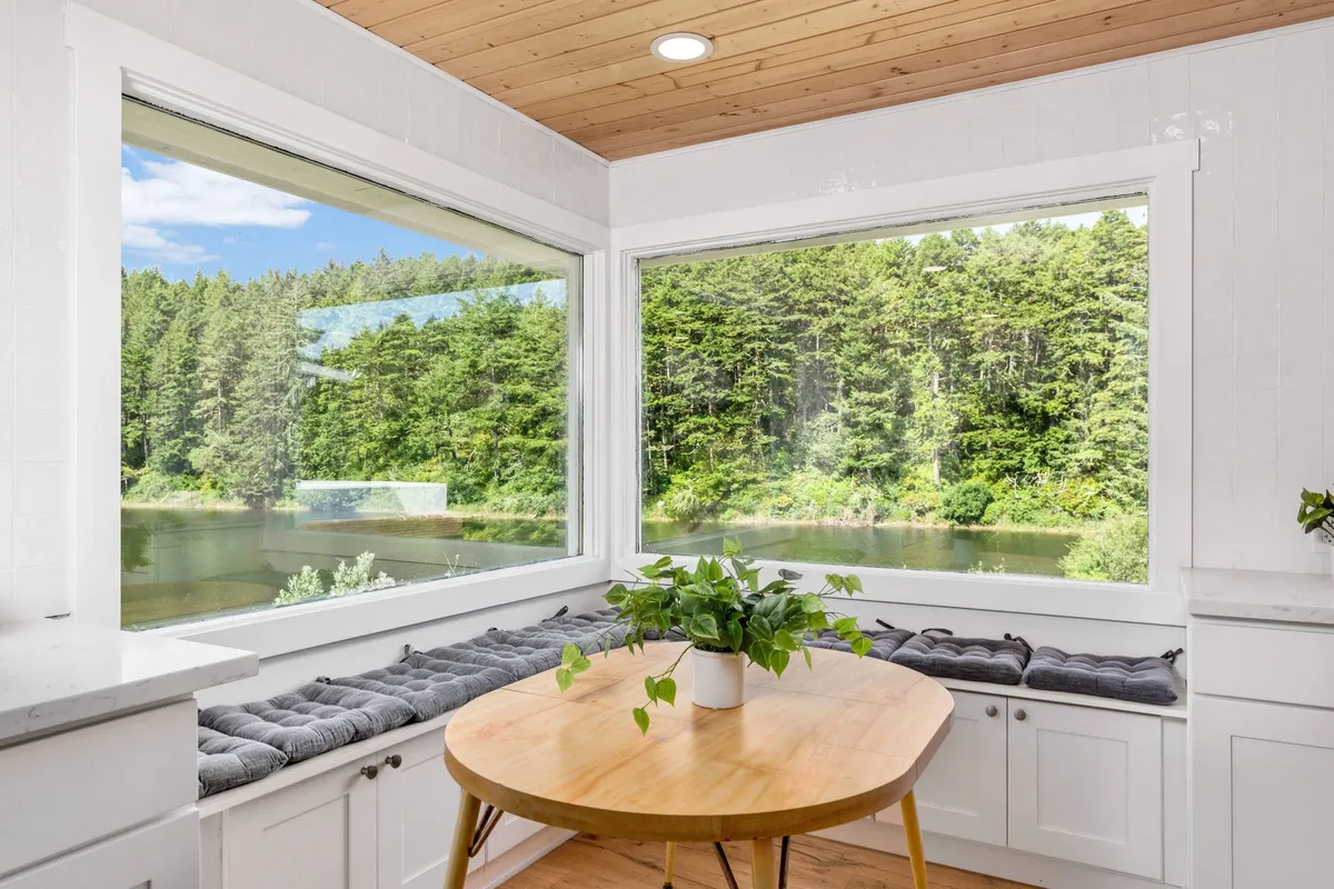 A serene dining nook featuring large windows overlooking the forest and water, a round wooden table, and comfortable window bench seating. The abundant natural light and expansive view connect the indoors to the natural beauty outside.