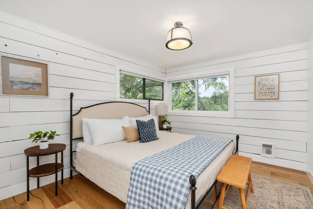 A serene bedroom with a single bed adorned with a blue checkered blanket, large windows letting in abundant natural light, and a small side table with a plant. The room feels cozy and tranquil, ideal for restful nights or quiet mornings.