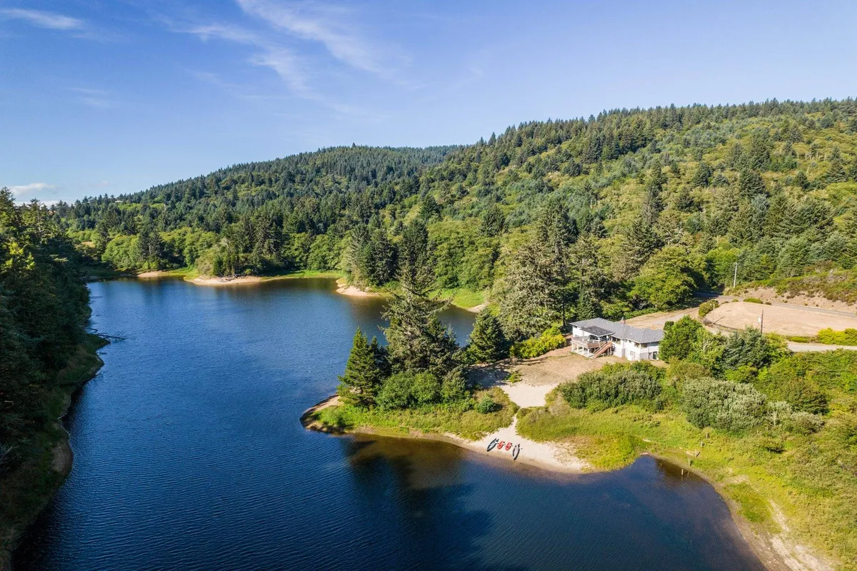 The image features a beautifully renovated lakefront house surrounded by lush trees, with a private sandy beach leading to the tranquil waters of Lake Legacy. The serene setting highlights opportunities for swimming, fishing, and enjoying sunset boat rides, while a cozy campfire area beckons for evening gatherings. The landscape is framed by the peacefulness of nature, offering both adventure and relaxation.
