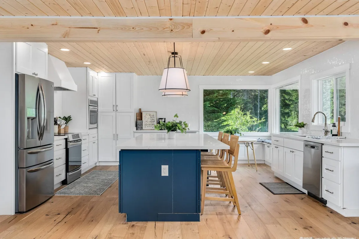 This image displays a spacious, inviting kitchen featuring a large kitchen island, bright windows letting in natural light, stainless steel appliances, and warm wooden beams overhead. The layout is perfect for group meal prep, casual chats, and family gatherings.