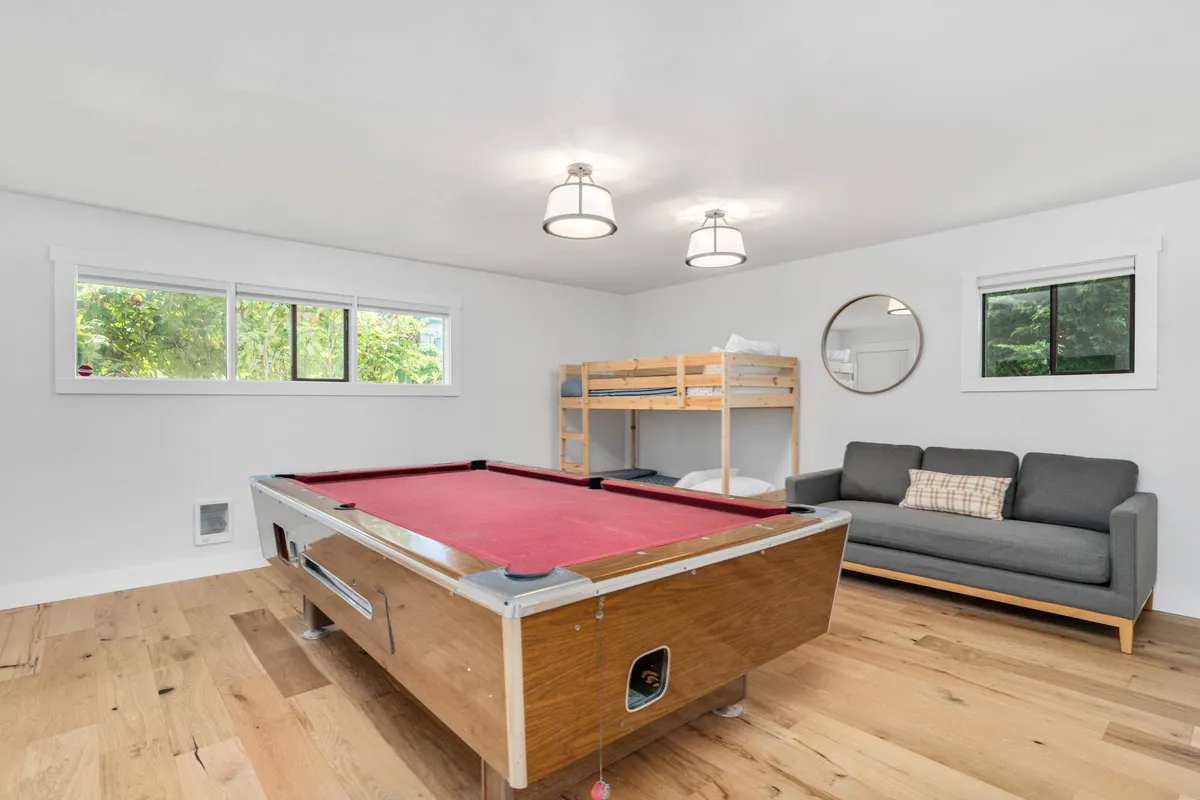 A playful, multifunctional room featuring a pool table, comfortable sofa, bunk bed for flexible sleeping arrangements, and large windows letting in natural light. The scene is set for both relaxation and fun family activities.