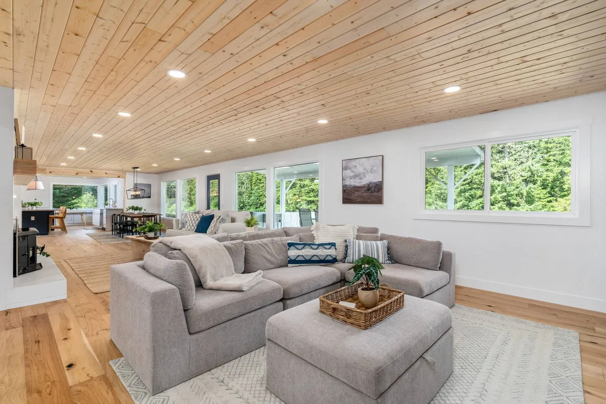 A large sectional sofa sits beneath a wooden ceiling, with a dining area visible in the background. The living space is framed by expansive windows overlooking lush greenery, giving the room a cozy-yet-open feel that encourages family gatherings and entertainment.