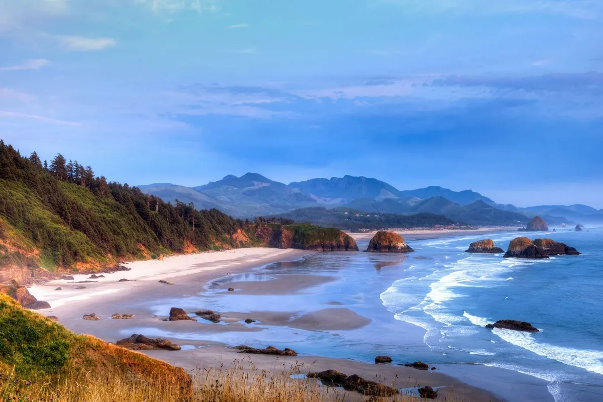A tranquil scene of an expansive beach bordered by unique coastal rocks, with gentle waves lapping the shore and distant mountains creating a picturesque horizon. The area looks ideal for leisurely walks, shell hunting, and soaking up the ocean breeze.
