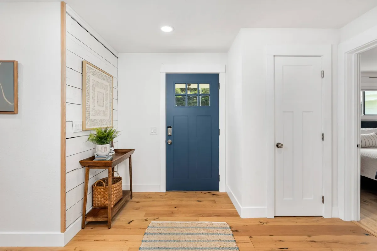 This image features a blue front door opening into a space with a wooden table, framed artwork, a woven basket, and a striped rug, all bathed in inviting natural light. The front entryway hints at the transition point between inside comfort and outdoor exploration.
